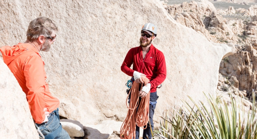 Standing amongst tall rock walls, two people appear to be speaking with each other, while one gathers climbing rope. 